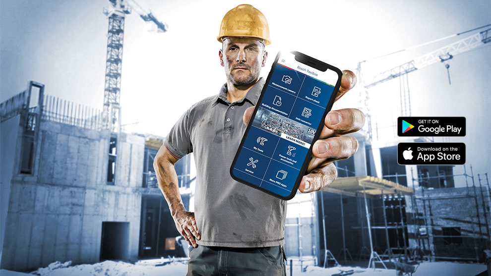 A construction worker wearing a helmet holds a smartphone in front of a construction site.
