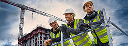 Three construction workers in reflective vests and helmets stand at a construction site.