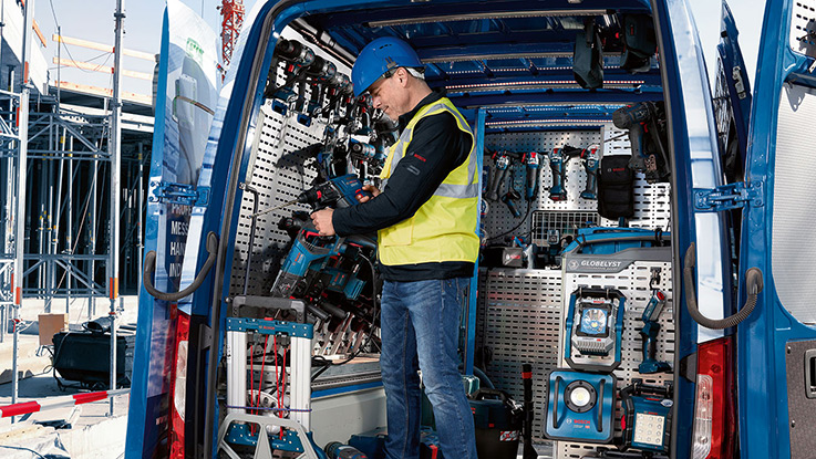 A craftsman in a yellow safety vest and blue helmet stands in a delivery van surrounded by tools.