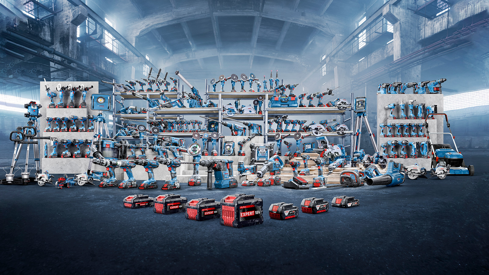 A variety of blue power tools and accessories on shelves in an industrial hall. The tools are organized, including cordless drills, saws, and bits. In the foreground, several red batteries are labeled "EXPERT 18V". The environment appears modern and well-lit, with an industrial flair.