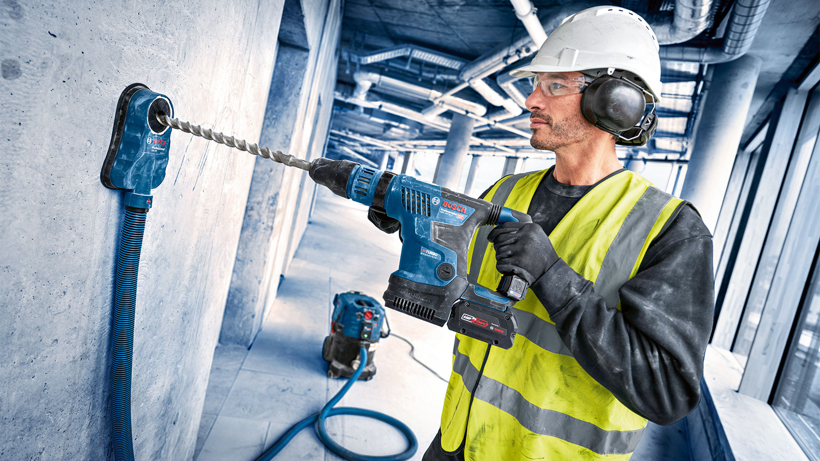 A man in a yellow safety vest and helmet is drilling with a blue Bosch Professional cordless drill into a concrete wall. Behind him, pipes and a construction site are visible.