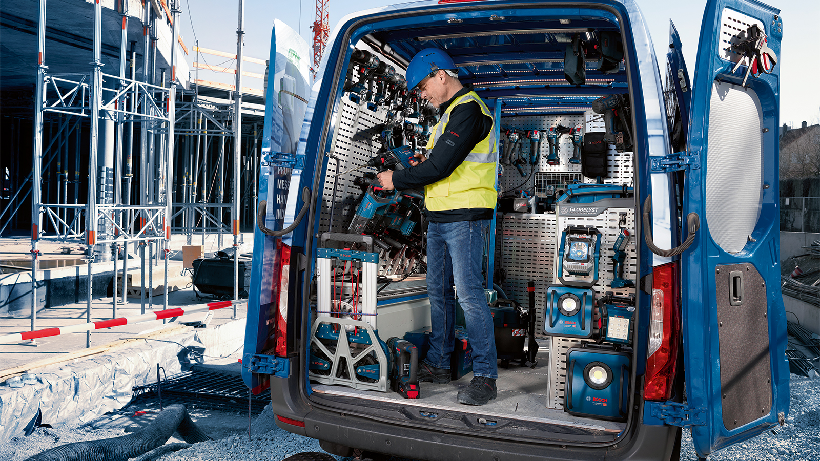 A craftsman in a yellow safety vest and blue hard hat stands in the open rear of a white van. Inside, various Bosch Professional tools are visible, including cordless drills and chargers, neatly arranged. In the background, construction scaffolding and a construction area can be seen, indicating an active work environment.