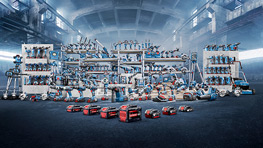 A wide selection of tools is displayed on shelves in an industrial hall. The tools are arranged in various colors, mainly blue and red. In the foreground, some red devices are on the floor. The hall has a gloomy atmosphere with light fog and high ceilings.