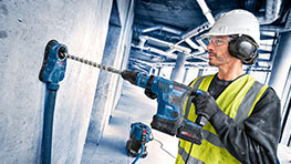A man wearing a helmet and safety vest is drilling into a wall with a Bosch drill.