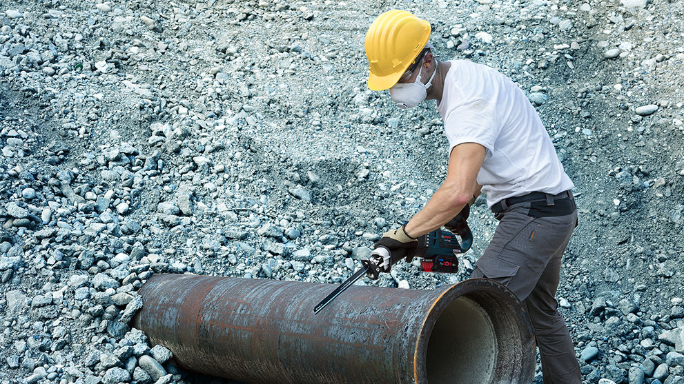 A worker with a yellow helmet is cutting a metal pipe on debris.