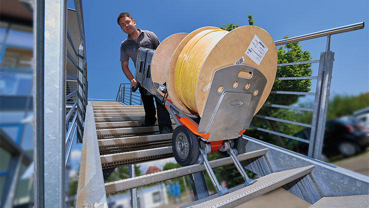 A man is pulling a hand truck with cable reels down the stairs.