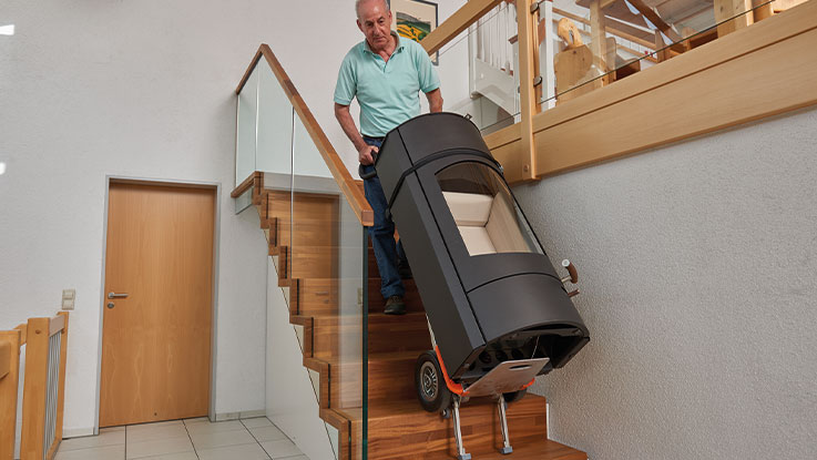 An older man is carrying a black stairlift down the stairs.