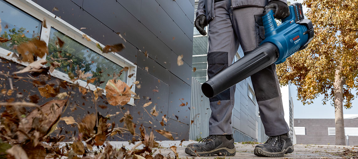 A worker is blowing leaves with a blue Bosch leaf blower in front of a building.