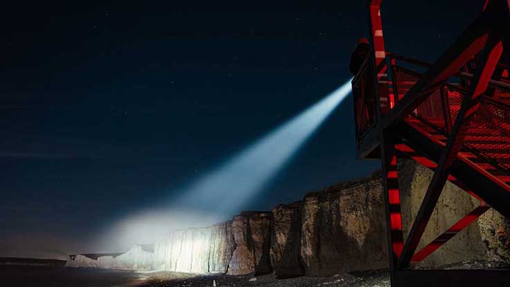 A red lighthouse shines light on cliffs and beach at night.
