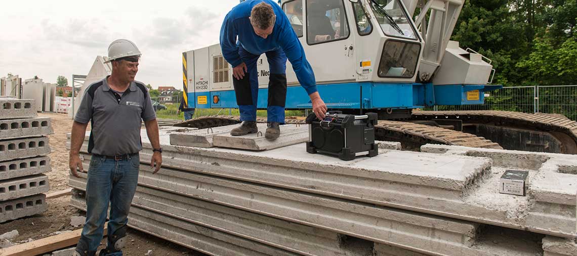 Two construction workers stand next to concrete slabs and a crane.