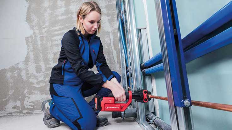 A woman in blue work clothes is cutting pipes with a red Bosch machine.