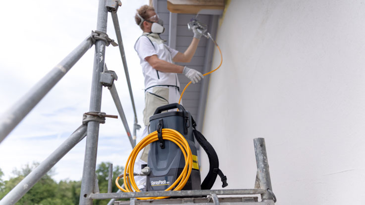 A craftsman is spraying paint on a wall, standing on a ladder.