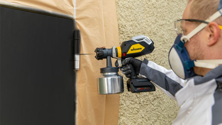 A craftsman is spraying paint on a wall with a Wagner spray gun.