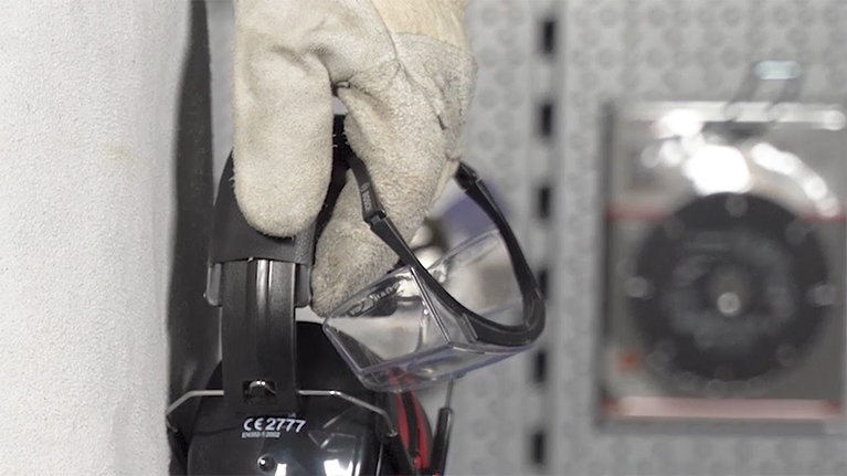 A hand in a white glove holds safety goggles on a hook.