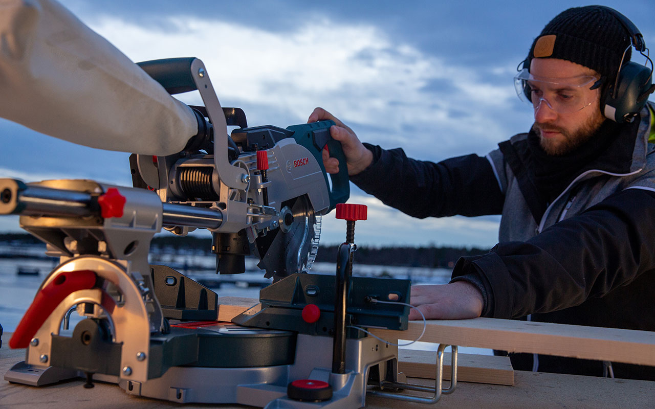 A man is cutting wood with a Bosch Professional miter saw at sunset.