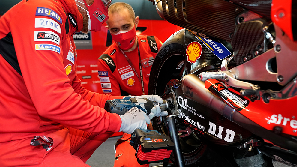 Two mechanics in red overalls are working on a Ducati motorcycle axle.