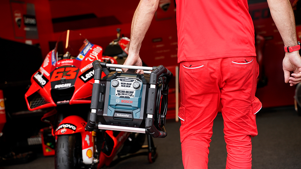 A man in red clothing is carrying a Bosch speaker next to a motorcycle.