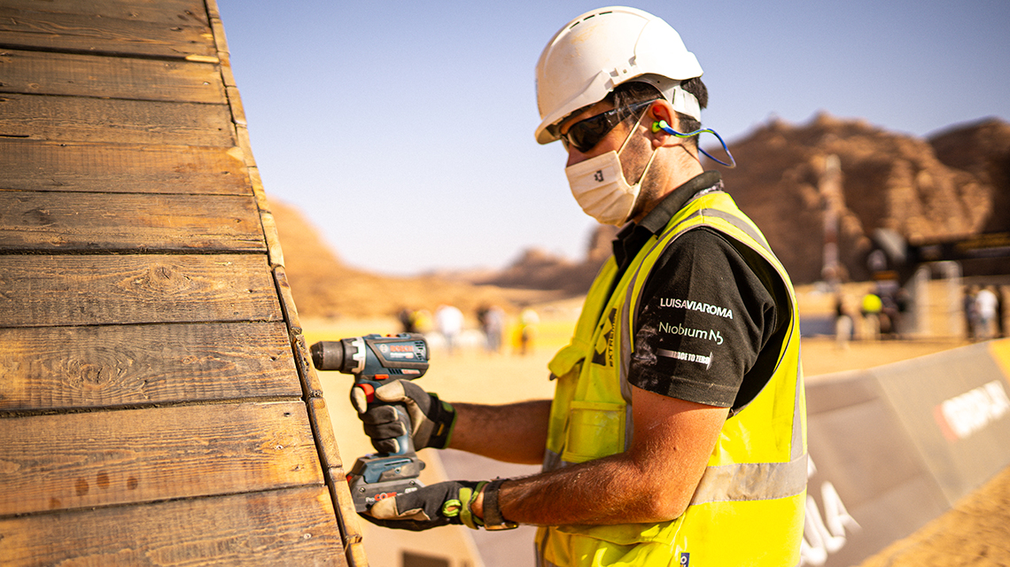 A worker with a cordless screwdriver at a wooden wall in the desert.