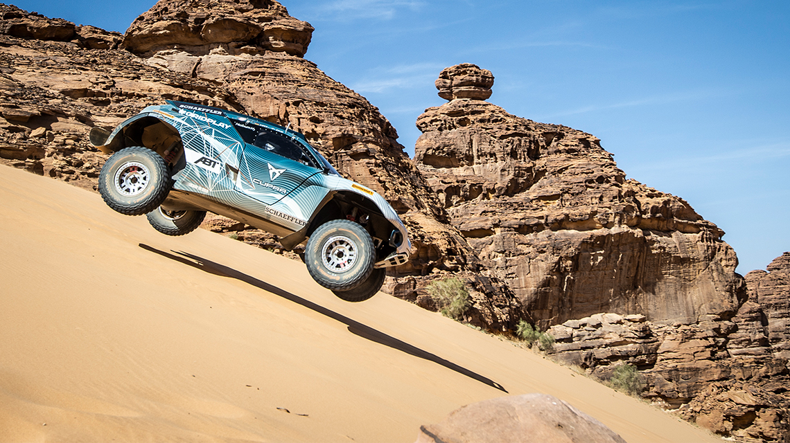 A blue off-road vehicle jumps over a sand dune in a desert.