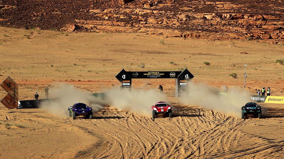 Three racing vehicles drive through dry sand, dust swirling up.