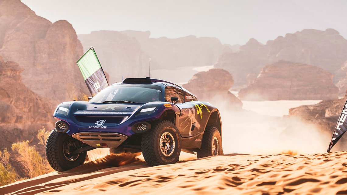 A blue off-road racing car drives through a sandy dune landscape.