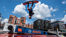 A wakeboarder jumps over a blue Bosch element in front of modern buildings.