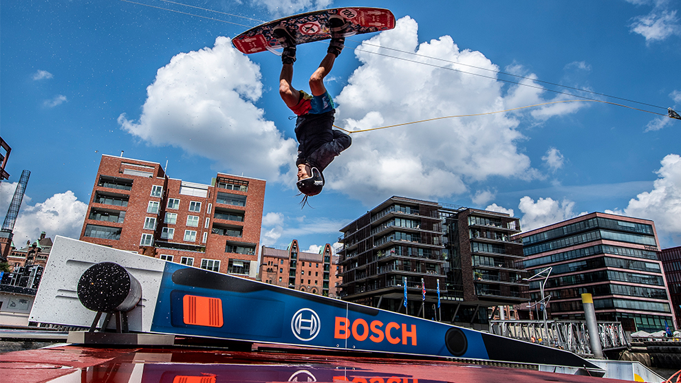 Wakeboarder jumps over a ramp, with modern buildings in the background.