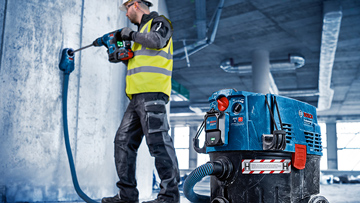 A worker in a yellow vest is drilling into a wall next to a blue vacuum cleaner.