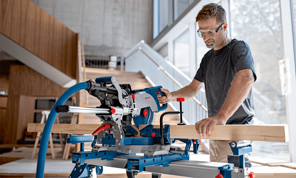 A man is working on a blue Bosch miter saw with wooden pieces.