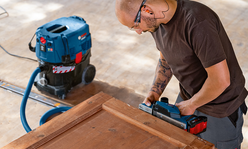 PLANING WOOD WITH PLANERS A man with glasses is working on wood with a blue Bosch tool.