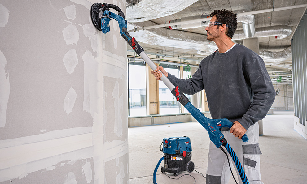 A craftsman with a Bosch sander is working on a wall in a room.