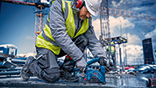 A construction worker in a yellow vest is using a blue Bosch saw on the construction site.