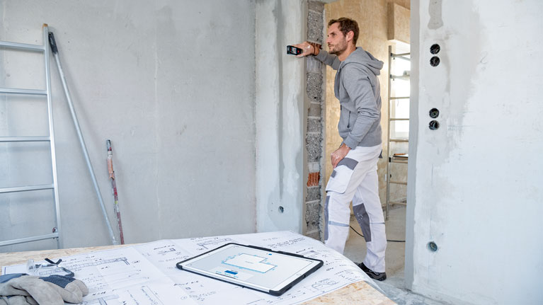 A craftsman measures with a laser on a wall, plans are next to him.