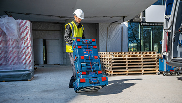 A worker in a yellow vest and helmet lifts a blue toolbox at the construction site. In the background, wooden pallets.