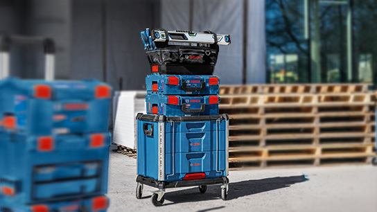 A stack of blue Bosch Professional toolboxes is on a wooden pallet, with a black bag of tools on top.