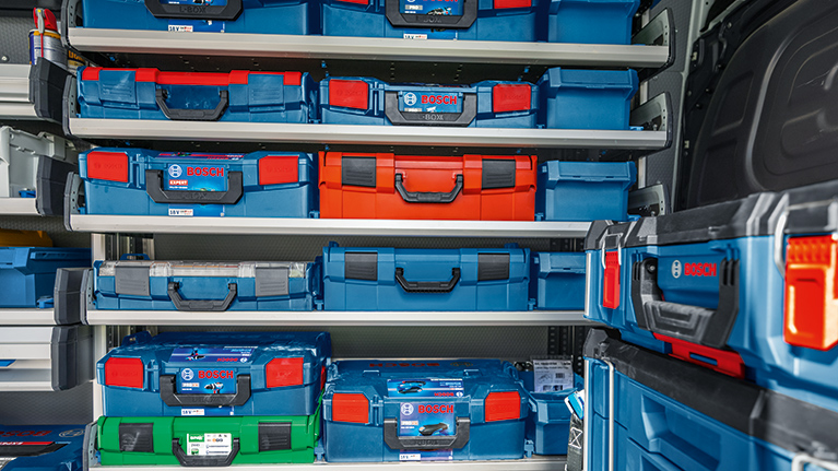 Several blue toolboxes from Bosch Professional are neatly arranged on shelves. The boxes come in various sizes, some featuring orange and green elements. The boxes are equipped with black handles and are located in a well-lit workshop.