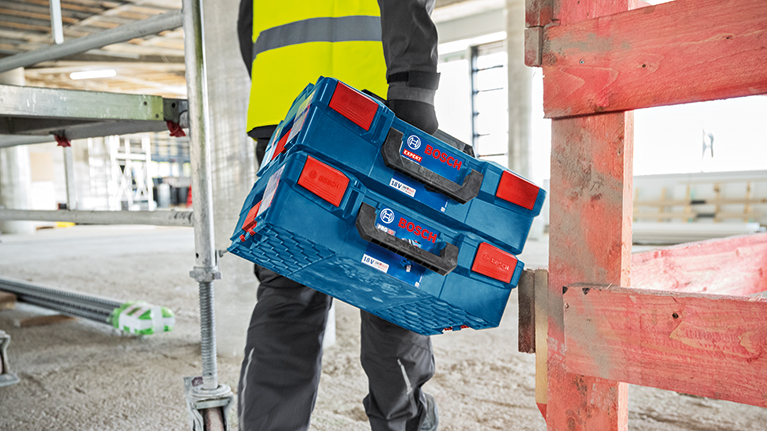 A worker in work clothes lifts a blue Bosch Professional toolbox with red handles onto another. The surroundings are bright and seem to be a construction site.