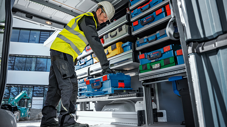 A worker in a yellow safety vest and helmet stands in a vehicle reaching for blue toolboxes. In the background, other colored boxes and a modern work environment are visible.