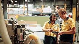 Two workers in yellow shirts are inspecting a machine in a workshop.