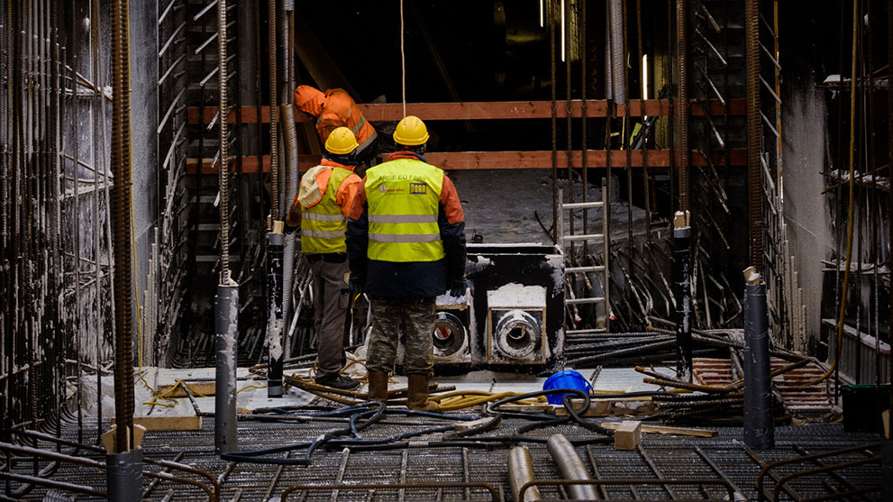 Three construction workers wearing helmets and vests at a construction site with pipes.