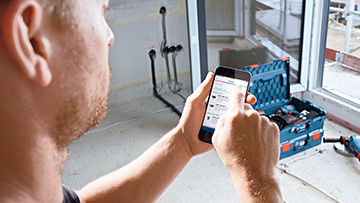 A man uses a smartphone in a room with a toolbox in the background.