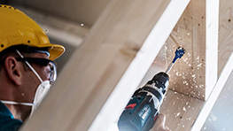 A craftsman with a hard hat is drilling into wood, shavings are flying around.