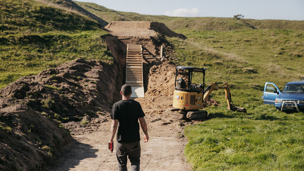 A man with tools is walking down a dirt path to a construction site with an excavator.