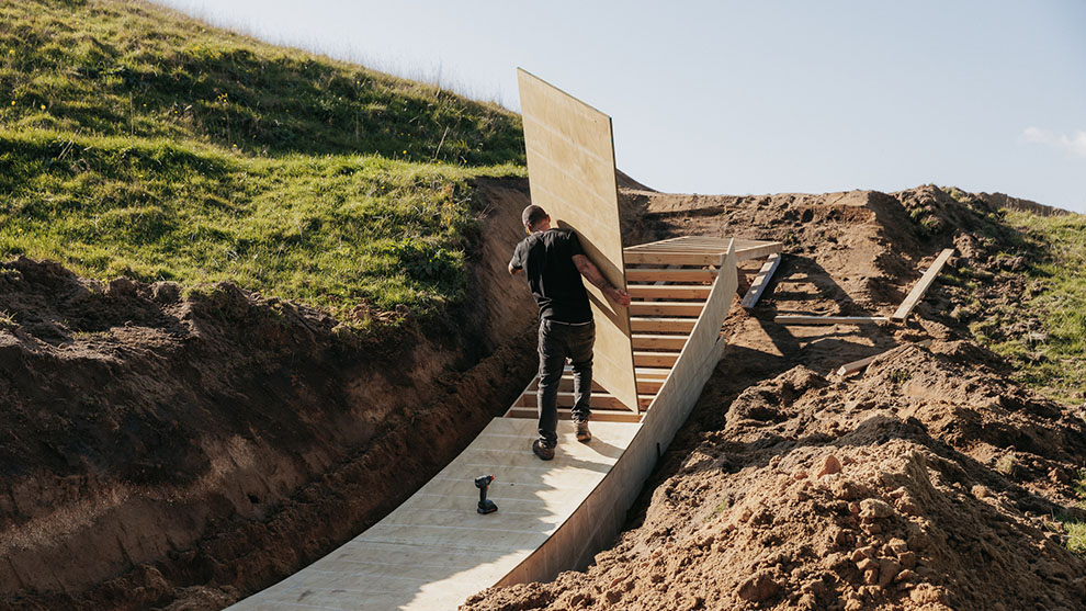 A man carries a large board on a wooden ramp in nature.