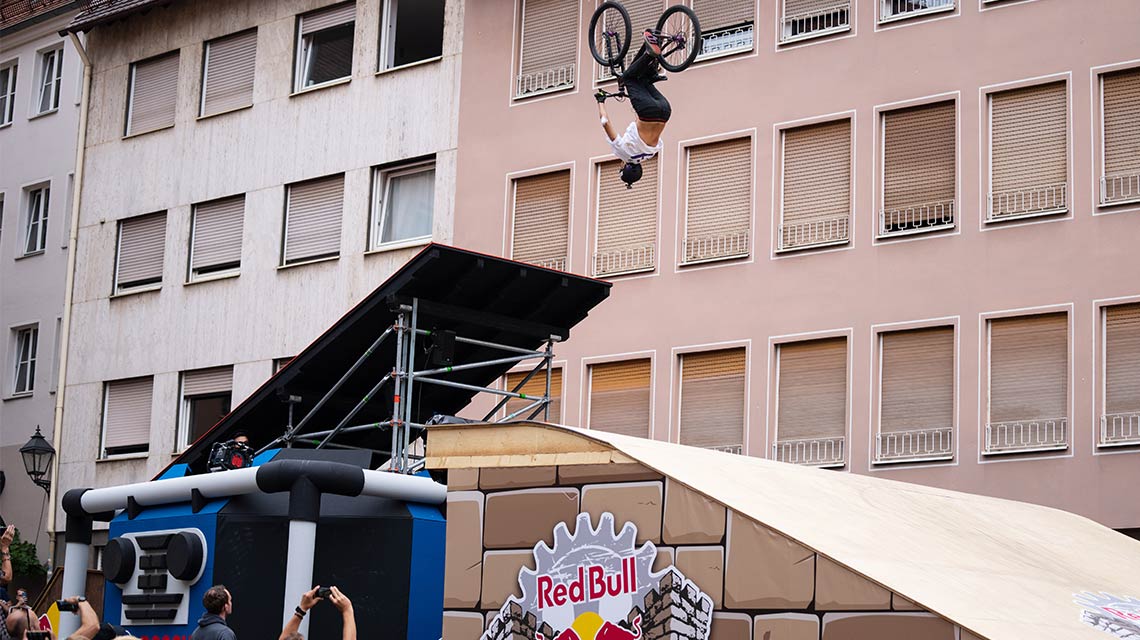 A BMX rider does a flip over a ramp in front of a building.