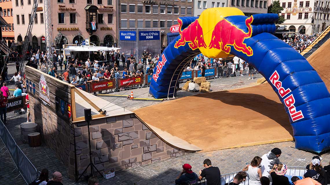 Colorful Red Bull jump gate over a dirt track, surrounded by spectators.