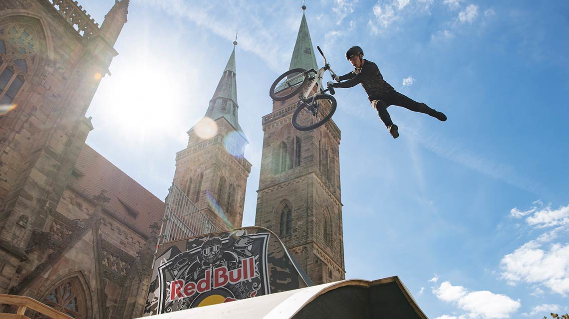 A cyclist jumps with a BMX over a Red Bull logo in front of churches.