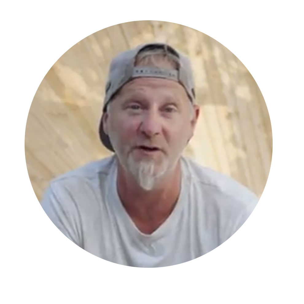 A man with short hair and a hat smiles in front of a wooden structure.