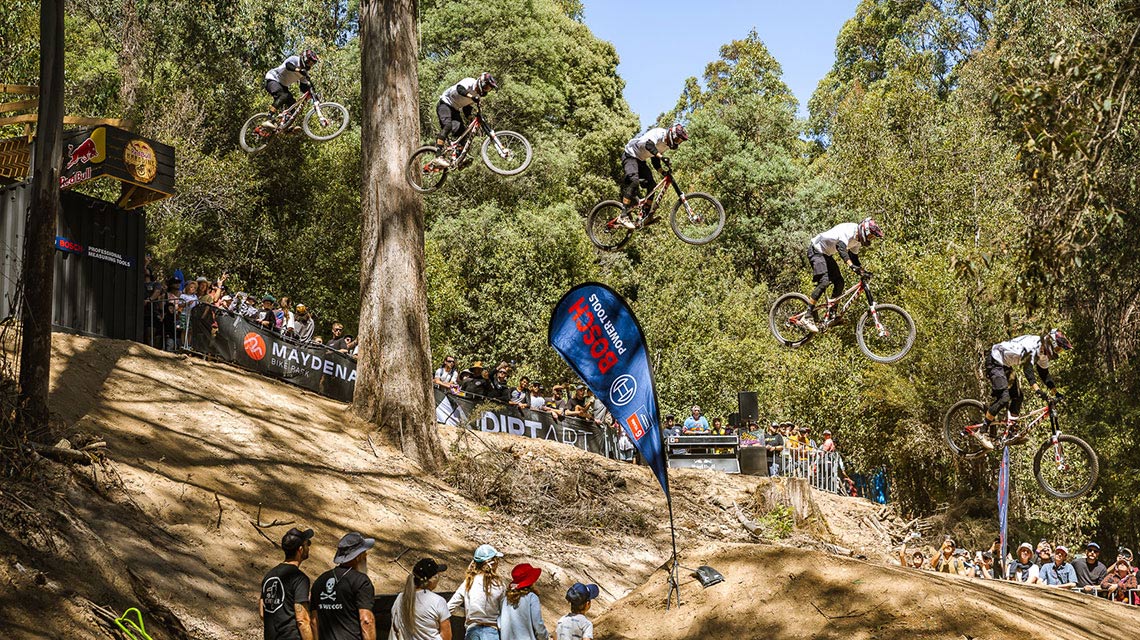 A cyclist jumps over a hill, spectators watch in the background.