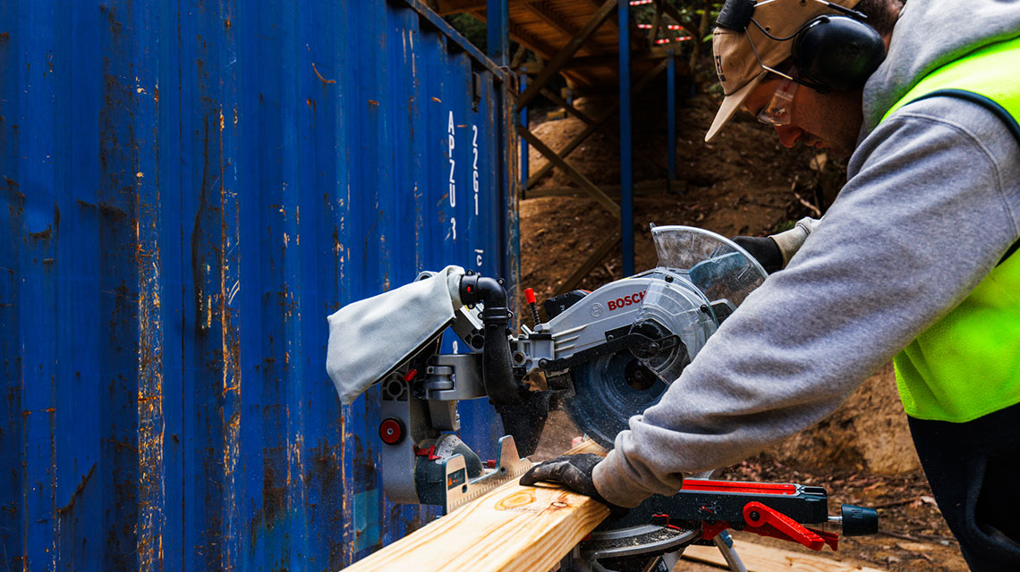 A craftsman wearing hearing protection is using a Bosch miter saw on a wooden board.
